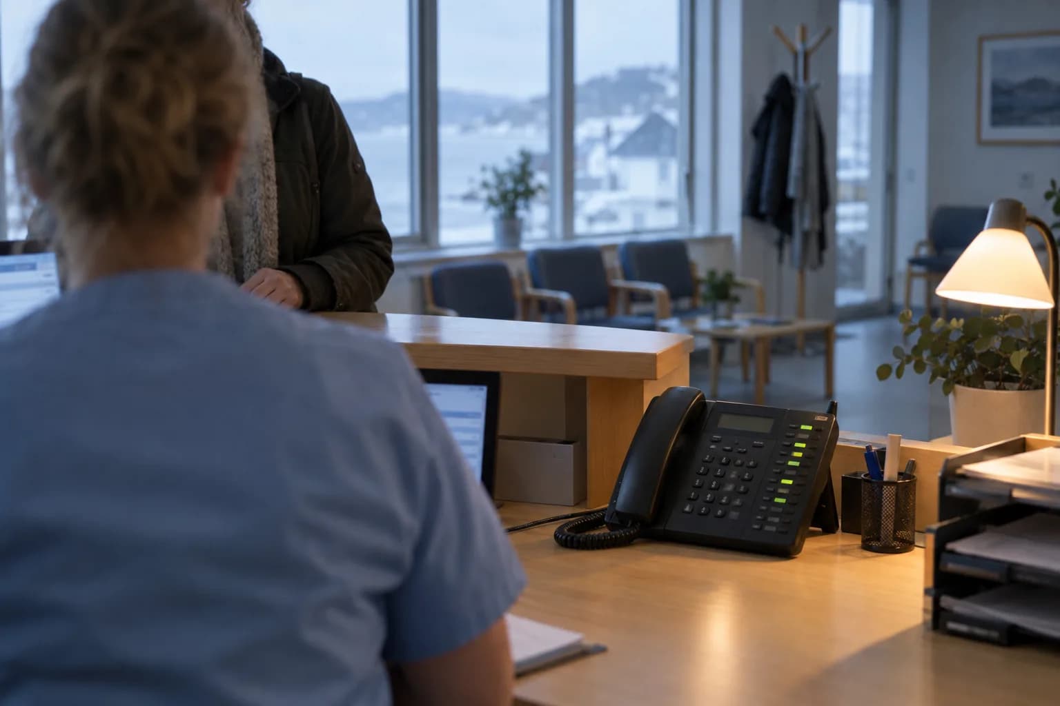 En helsesekretær i skranken på et legekontor hjelper en pasient, mens linjeknappene på bordtelefonen lyser