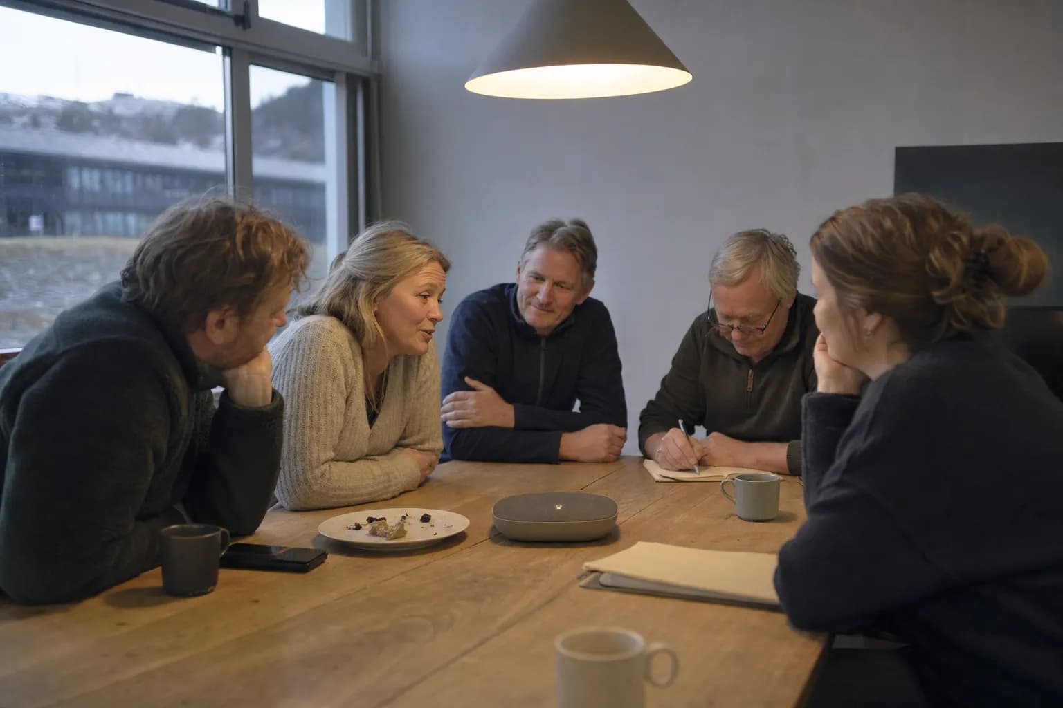 Fem kolleger sitter tett rundt enden av et møtebord og snakker mot en konferansetelefon midt på bordet, vendt mot den som mot en sjette kollega.