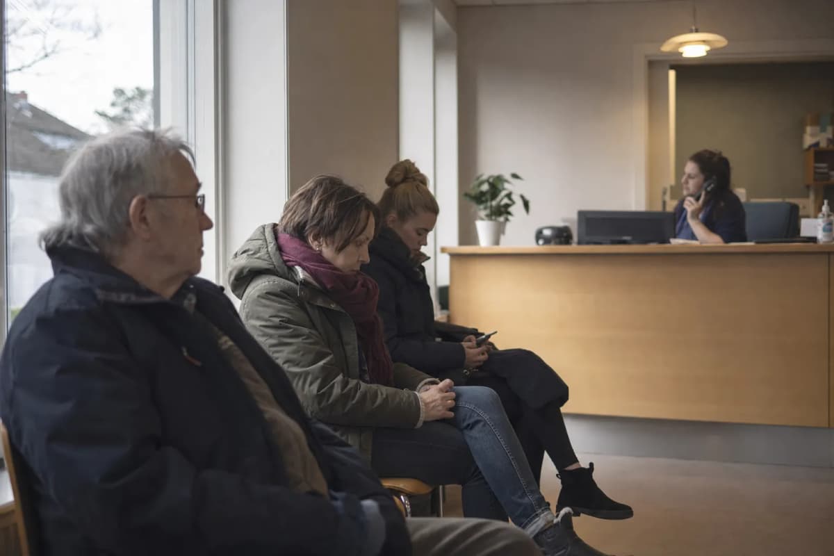 Venteværelse i en lægepraksis med patienter i stolene og en bemandet skranke