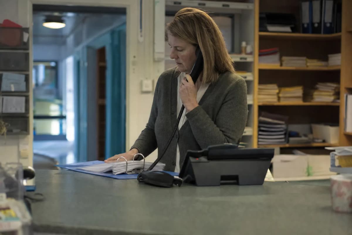 Medarbejder på et skolekontor ved en skranke med en telefon og en mappe