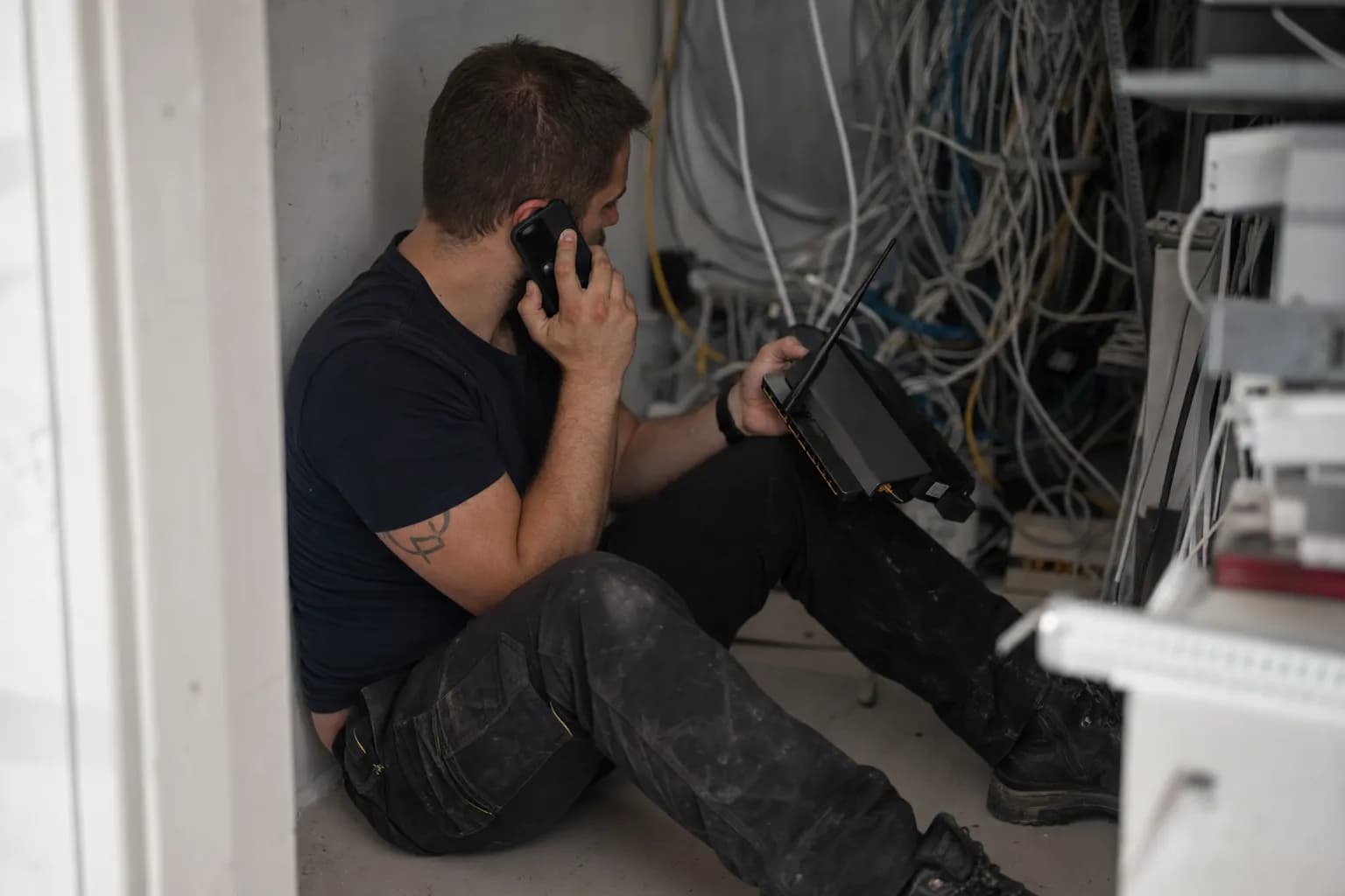 En tekniker på gulvet i et teknisk rom med en ruter i hånda og mobilen mot øret