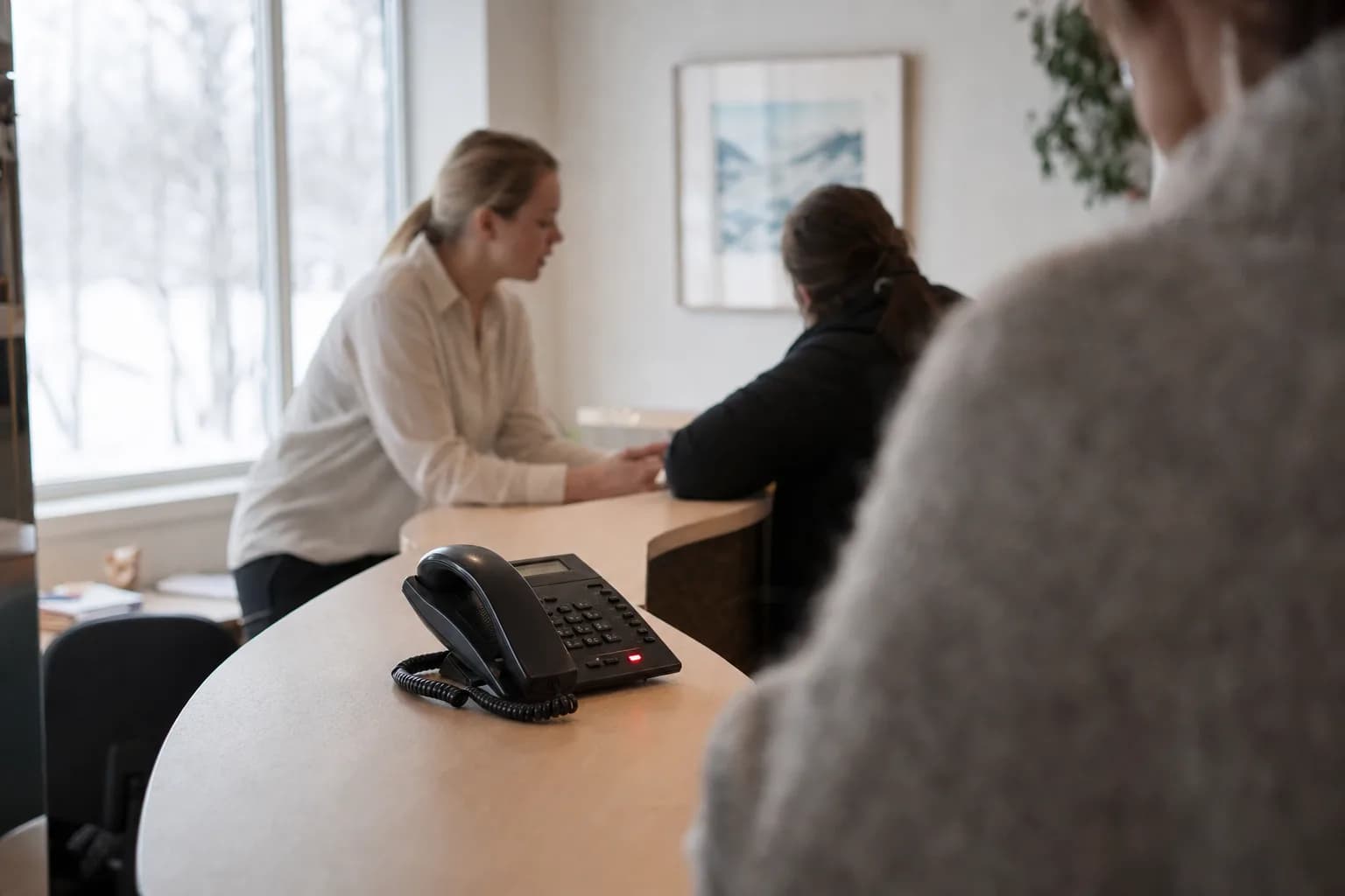 An office desk phone with a lit red message light stands on a pale clinic reception counter, while a receptionist in the background talks with a patient by a window with snow outside.