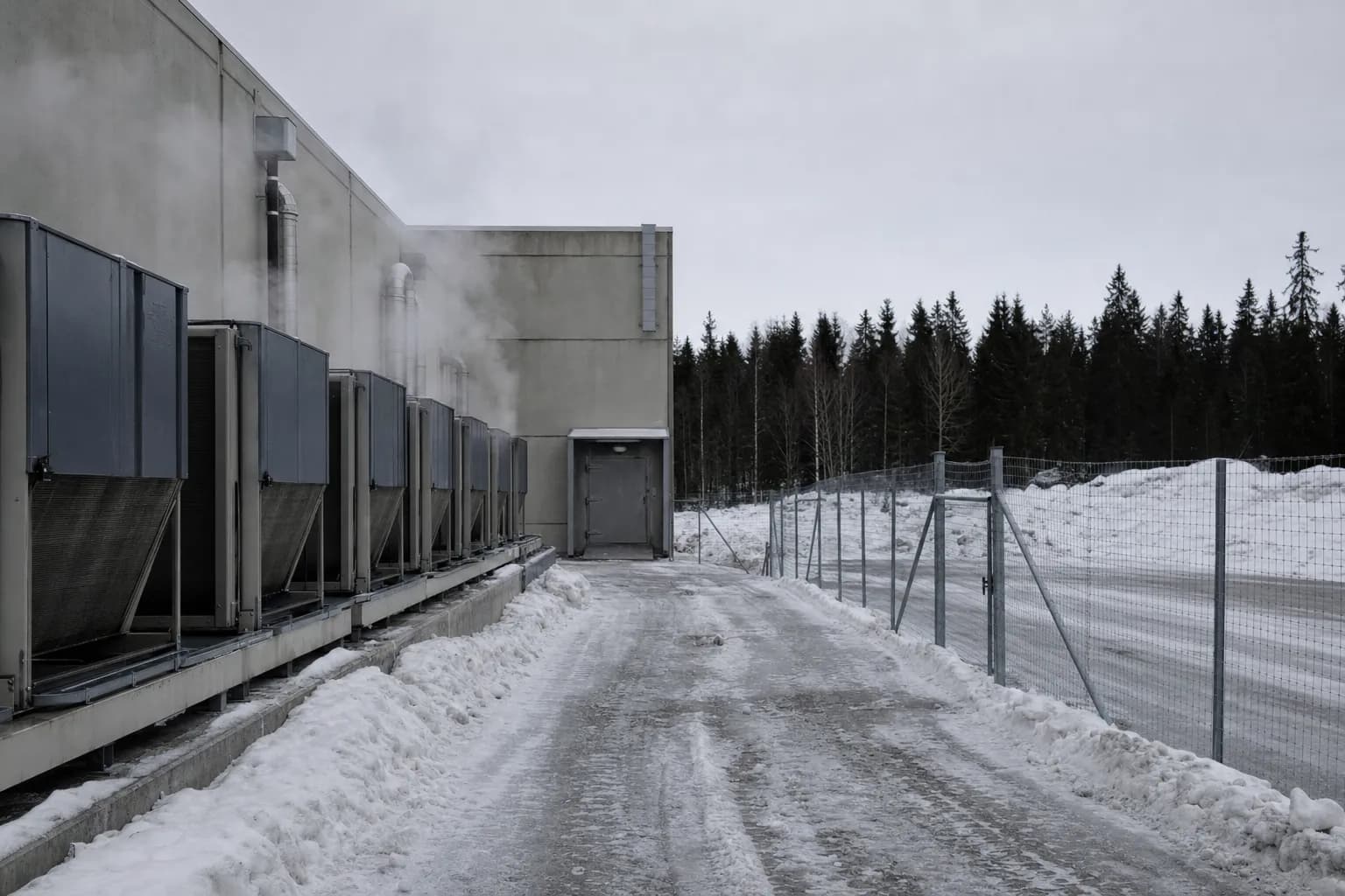 A low concrete building in a snow-covered Nordic industrial estate, cooling units along the wall