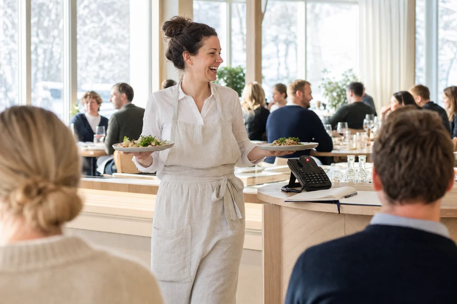 A smiling server carries two plates through a bright dining room with full tables and snow outside the windows. On the counter beside her a desk phone glows red with an unanswered call, next to an open reservations book.