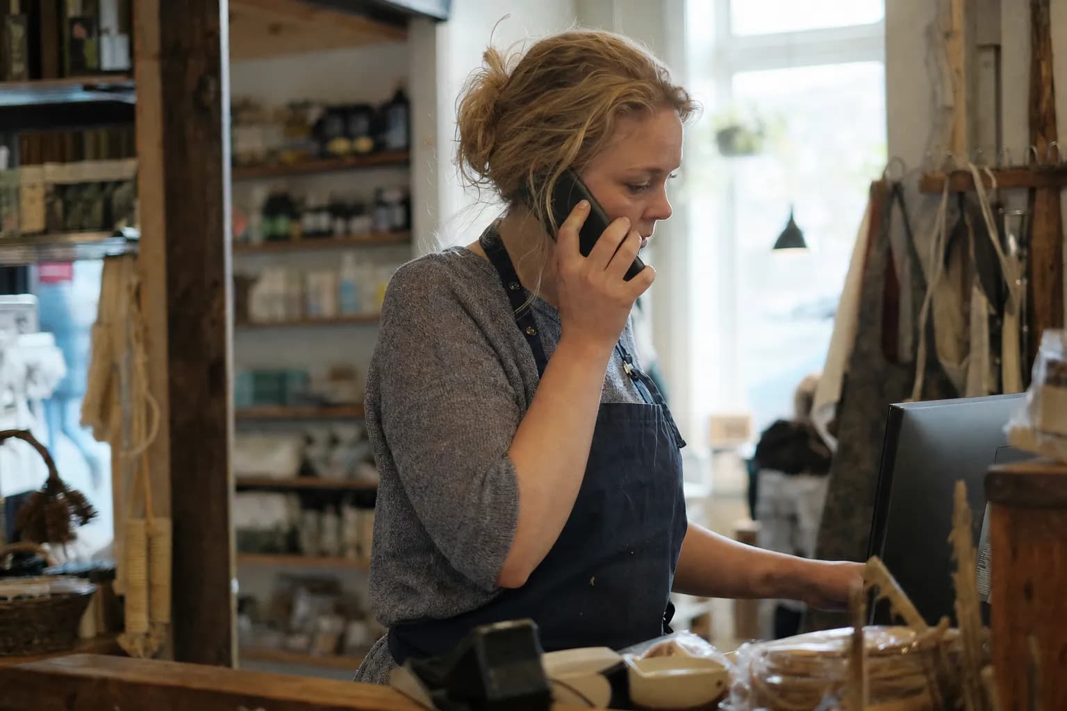 A shopkeeper behind the counter of a small shop with a phone to her ear