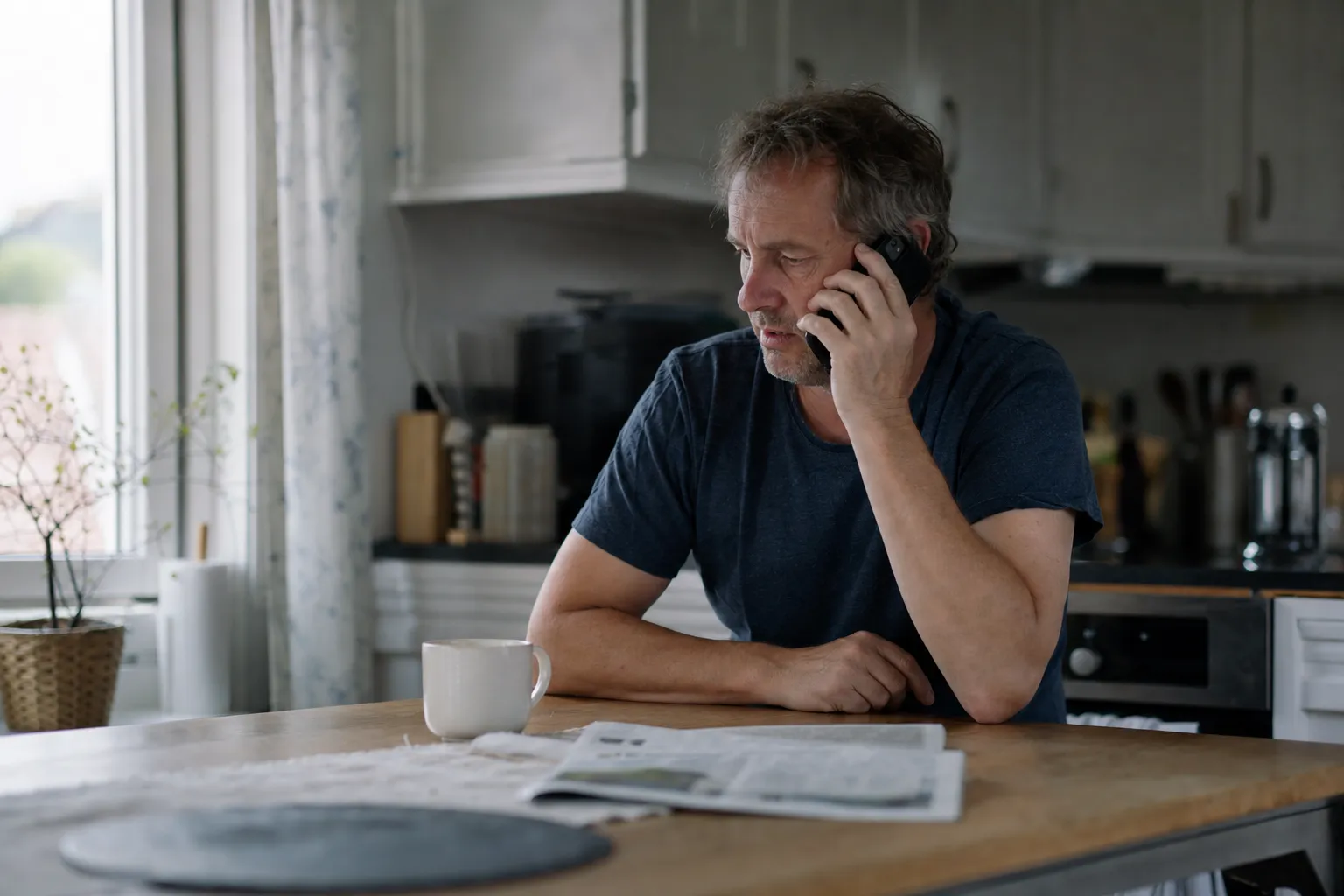 A man sits at his kitchen table at home with his phone to his ear