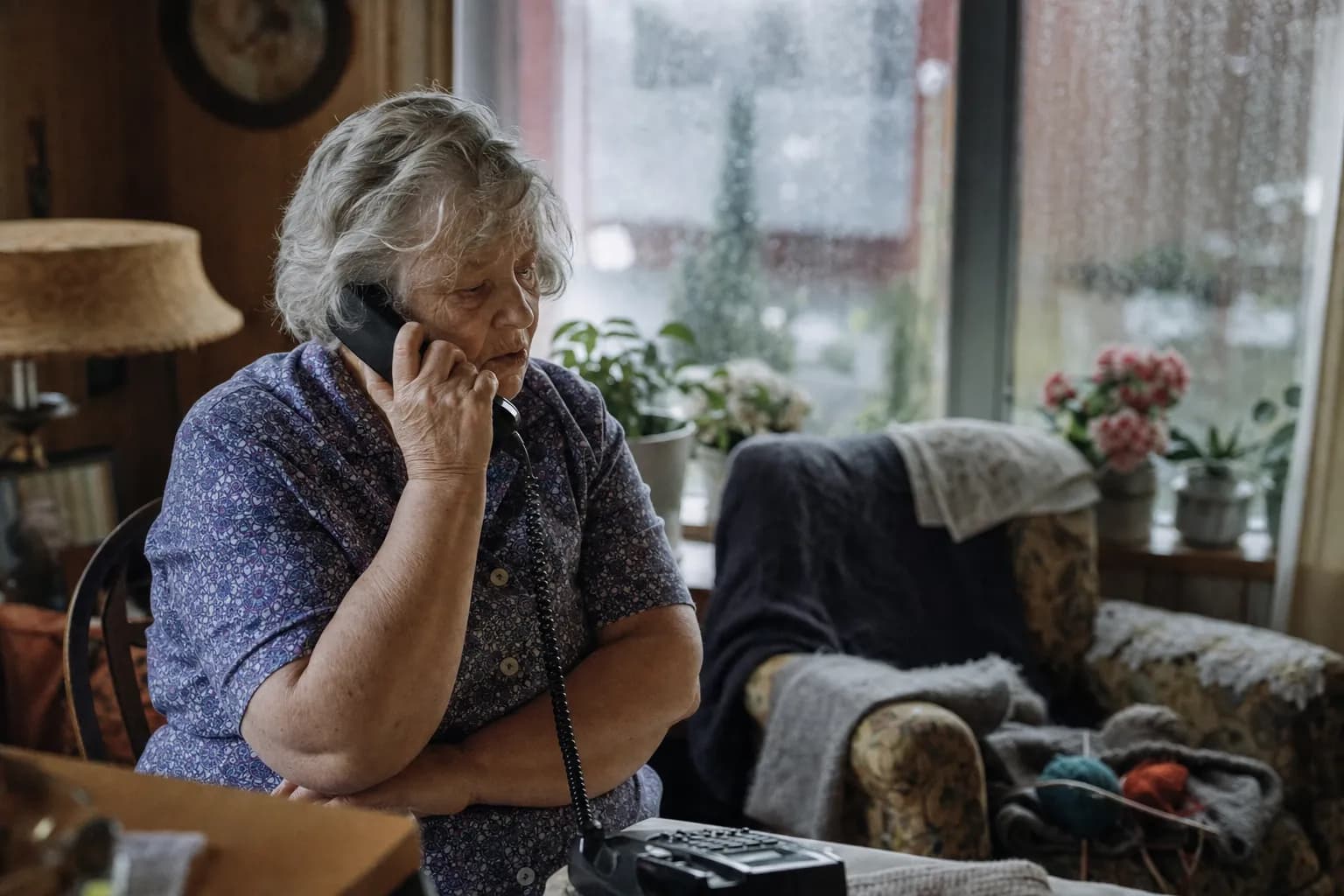 An older woman talks on a landline in a living room with rain on the window