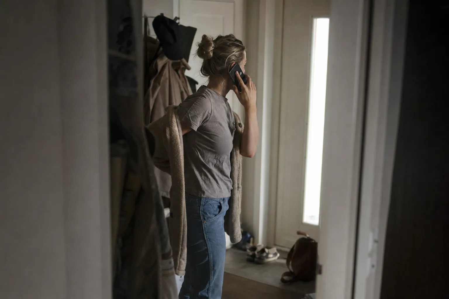 A woman stands in a hallway with her phone to her ear and her coat half on