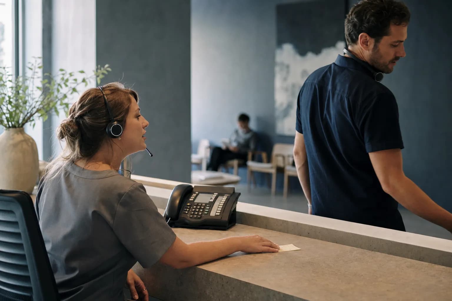 A woman in a headset sits at a clinic reception desk and starts to speak, while a colleague who has just placed a handwritten note on the counter beside her is already walking away