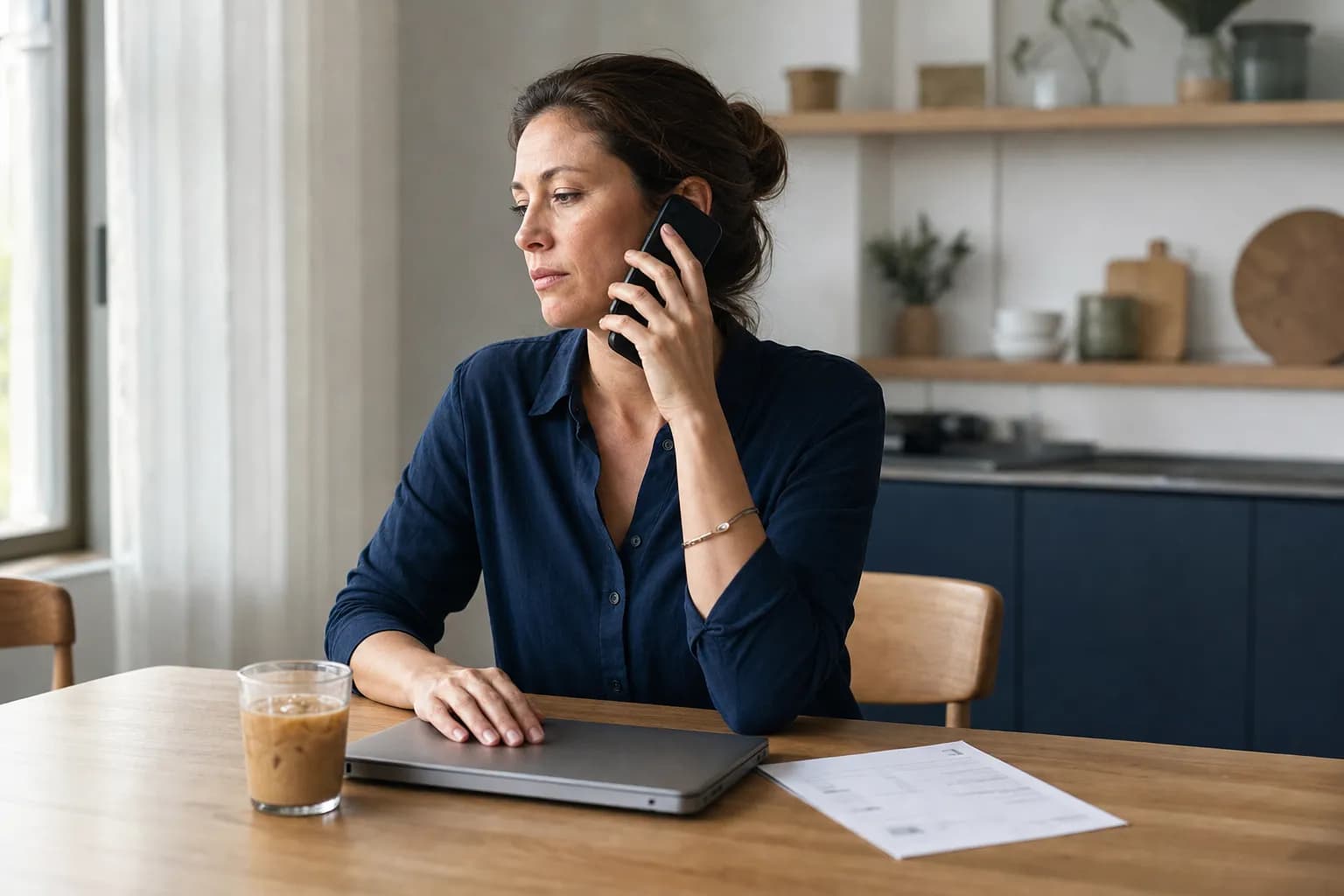 Woman sitting at a kitchen table in a bright apartment with a mobile phone to her ear, one hand resting on a closed laptop and an invoice lying on the table beside her