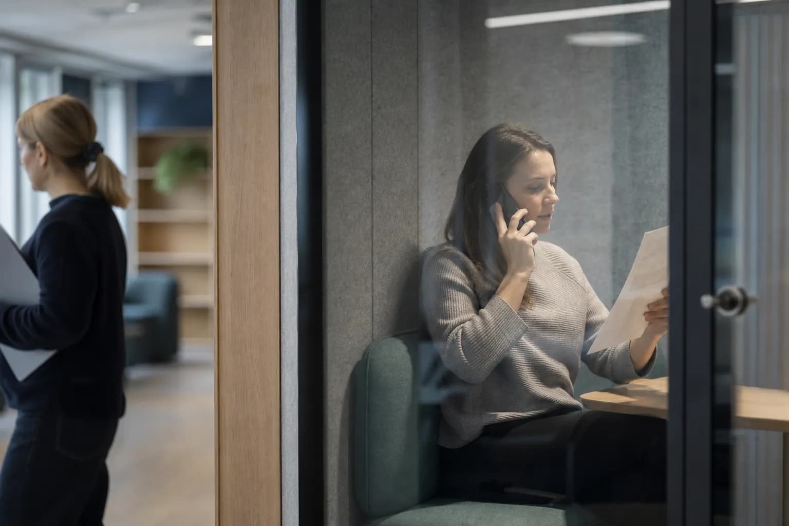 Seen through the glass of a phone booth in a co-working space, a woman stands mid-call with a mobile to her ear and a single printed sheet held up in her other hand, while a colleague waits out of focus in the foreground