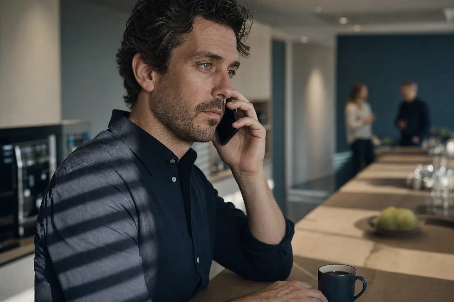 Close-up of a man in a dark shirt standing at an office staff kitchen counter, listening with a mobile phone to his ear, afternoon light through the blinds striping his cheek while two colleagues talk out of focus behind him