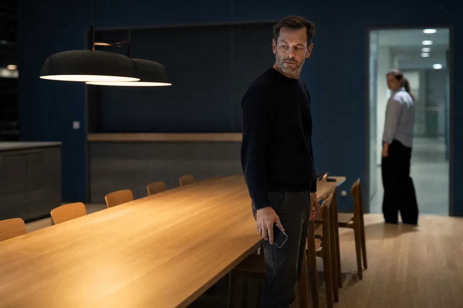 A man in a dark jumper stands by a long table in an empty staff canteen after hours, lit by two pendant lamps, having just lowered his phone to his side, while a woman in a light shirt has stopped in a lit doorway behind him