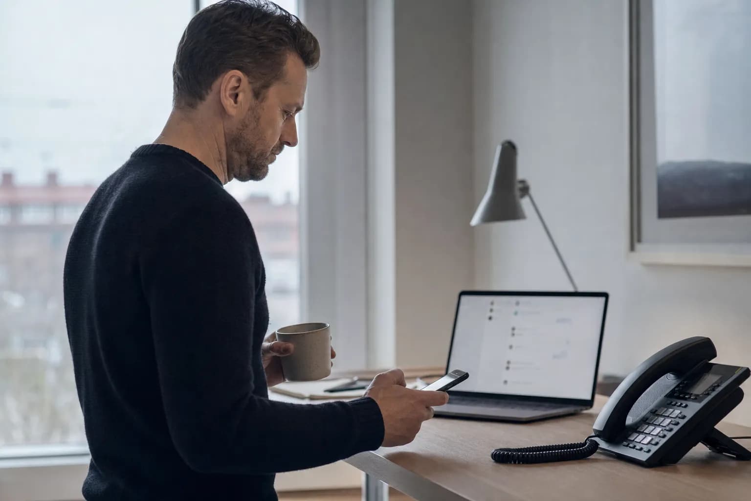 A man in a dark knit sweater stands at a standing desk in morning light holding a coffee mug in one hand and a phone in the other, with an open laptop and a matte black desk phone with a coiled cord on the desk in front of him