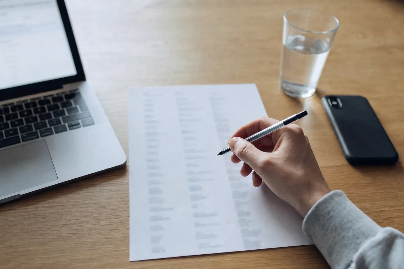 A desk seen from above with a printed list on white paper in the middle, a hand resting on the page holding a ballpoint pen just above it, the edge of an open laptop to the left and a phone face down to the right
