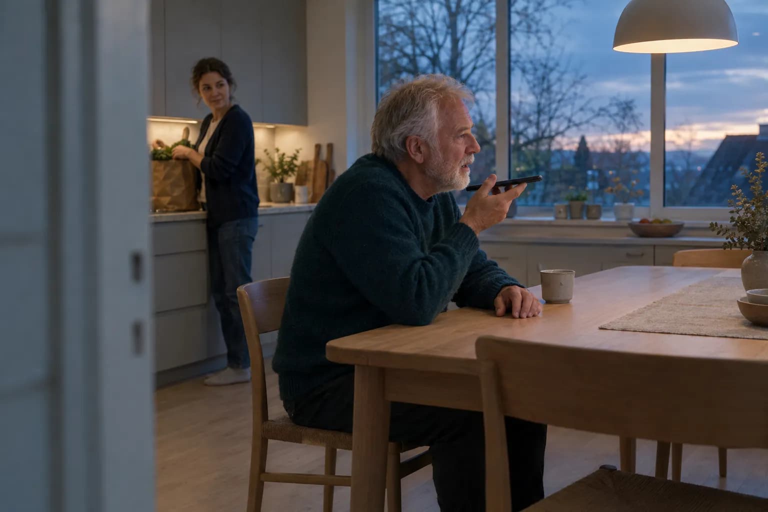 Eldre mann med hvitt skjegg i mørk blågrønn genser sitter ved et kjøkkenbord i eik i skumringen og snakker inn i mobilen han holder foran munnen, mens en kvinne i mørkeblå cardigan pakker ut en papirpose ved kjøkkenbenken bak og ser mot ham